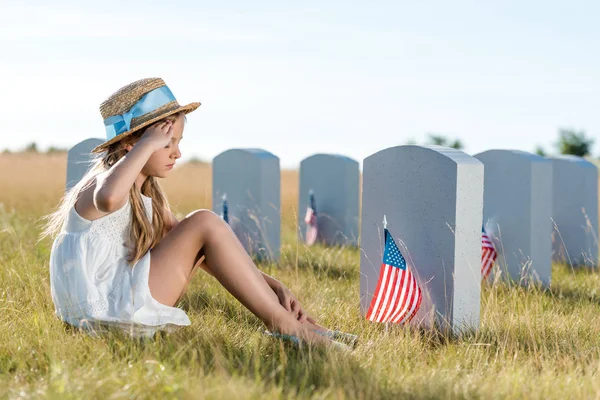 Kid in straw hat giving salute while sitting near headstones with american flags — Stock Photo