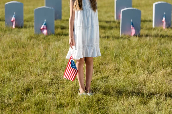 Ausgeschnittene Ansicht eines Kindes in weißem Kleid, das mit amerikanischer Flagge auf dem Friedhof steht — Stockfoto