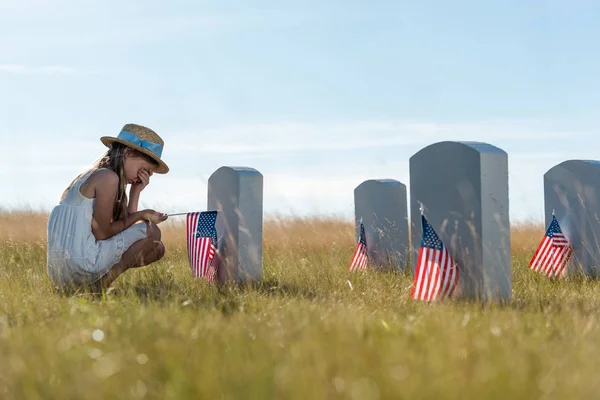 Selektiver Fokus des Kindes, das Gesicht verdeckt, während es in der Nähe von Grabsteinen mit amerikanischen Flaggen sitzt — Stockfoto