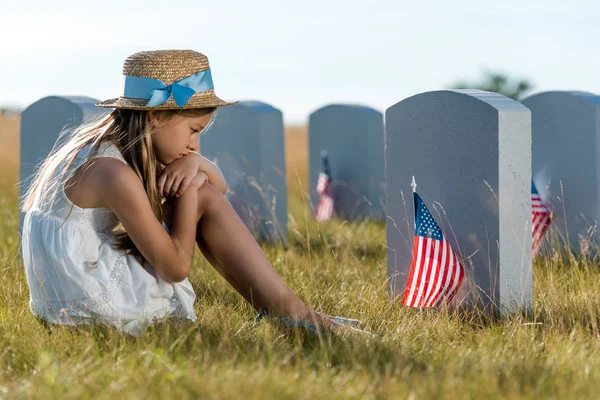 Selective focus of sad child sitting and looking at headstones with american flags — Stock Photo