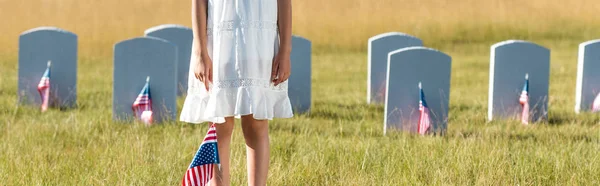 Panoramic shot of kid in white dress standing on graveyard with american flag — Stock Photo