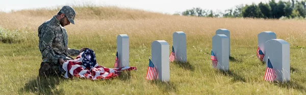 Tiro panorámico de triste soldado en uniforme de camuflaje y gorra con bandera americana mientras está sentado en el cementerio - foto de stock