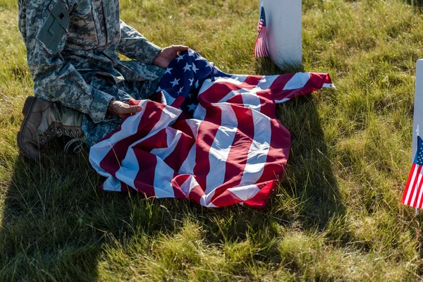 Vista recortada del hombre en uniforme de camuflaje con bandera americana y sentado en el cementerio - foto de stock