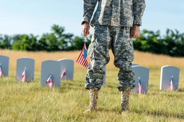 Vista recortada de soldado en uniforme de camuflaje sosteniendo bandera americana y de pie en el cementerio - foto de stock