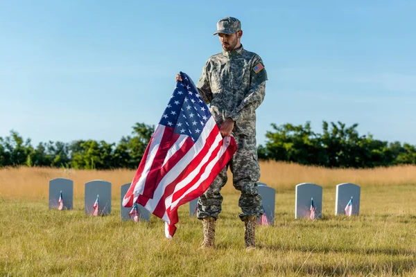 Apuesto veterano en uniforme de camuflaje sosteniendo bandera americana y de pie en el cementerio - foto de stock
