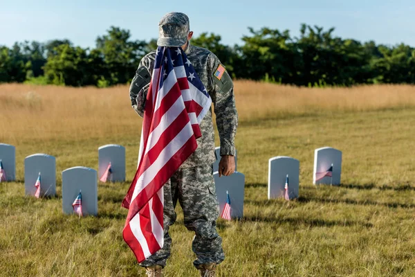 Soldat in Tarnuniform verdeckt Gesicht mit amerikanischer Flagge und steht auf Friedhof — Stockfoto