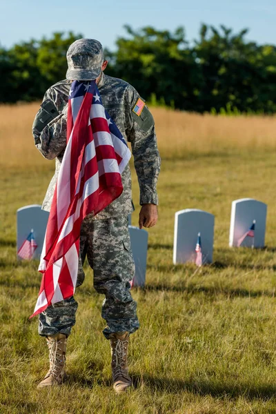 Homem de camuflagem uniforme cobrindo rosto com bandeira americana e de pé no cemitério — Fotografia de Stock