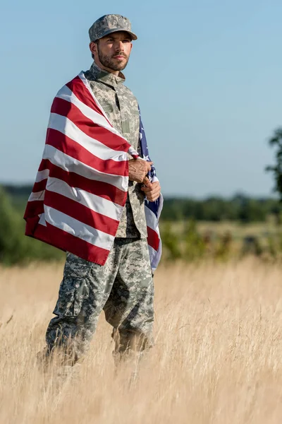 Homem em uniforme de camuflagem e boné segurando bandeira americana em campo dourado — Fotografia de Stock