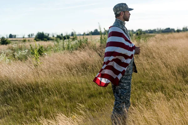 Vista lateral del soldado en uniforme de camuflaje con bandera americana en el campo de oro - foto de stock