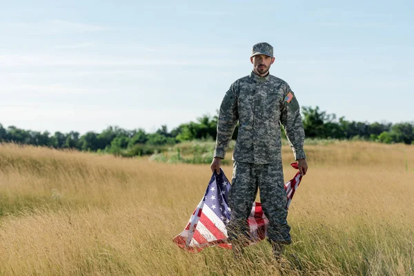 Soldado bonito em uniforme de camuflagem segurando bandeira americana no campo dourado — Fotografia de Stock