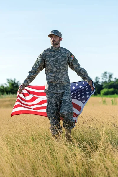 Guapo soldado en uniforme de camuflaje con bandera americana en el campo - foto de stock