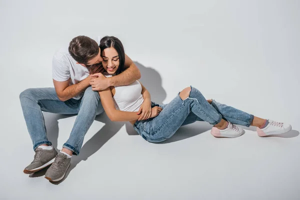 Повний вигляд молодого чоловіка цілує дівчину, коли вона посміхається і сидить на підлозі — стокове фото