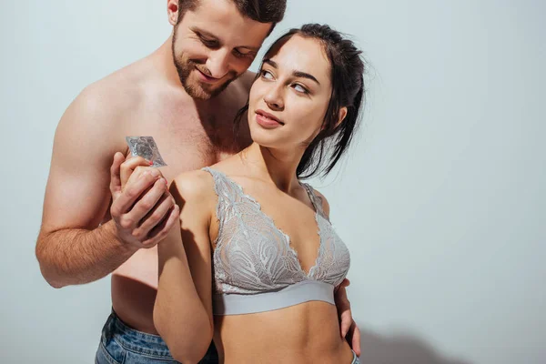 Сексуальная пара улыбается и обнимается, пока девушка держит презерватив и смотрит на мужчину — стоковое фото