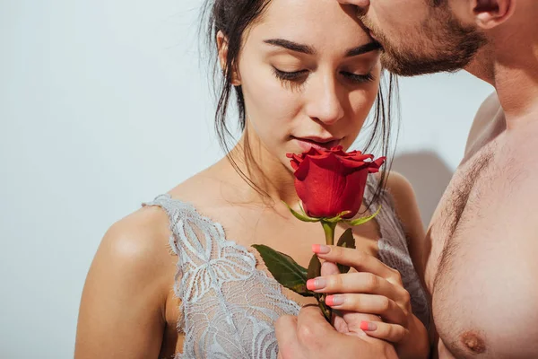 Guapo hombre desnudo besando novia en la frente mientras chica oliendo rosa roja - foto de stock