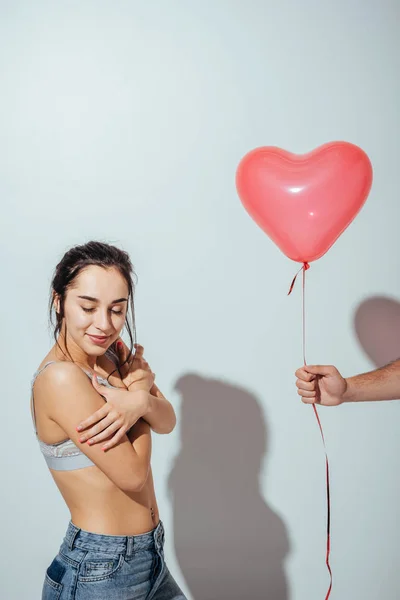 Обрезанный вид человека, дающего красный шар в форме сердца привлекательной девушке, которая стоит с закрытыми глазами — стоковое фото