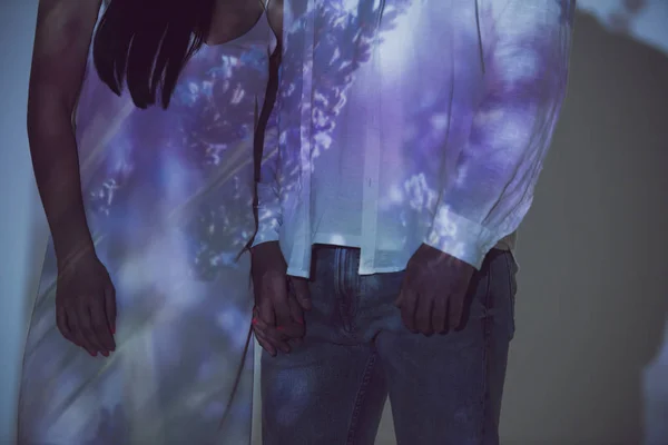 Cropped view of couple standing near each other in darkness in light and holding hands — Stock Photo