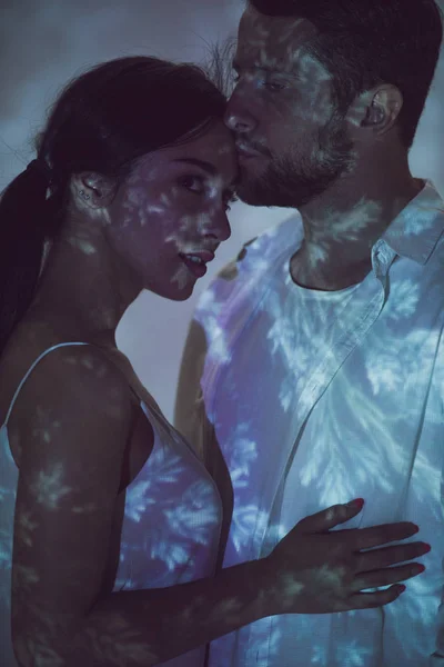 Couple in love standing in darkness and hugging while man kissing forehead of girl — Stock Photo