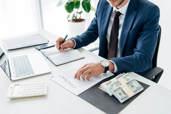 Vista recortada del hombre en traje de escribir en cuaderno cerca de dinero y portátil - foto de stock