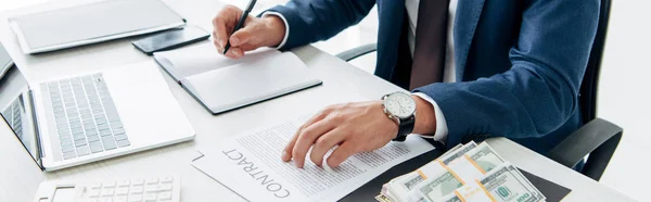 Panoramic shot of man in suit writing in notebook near laptop, money and contract — Stock Photo