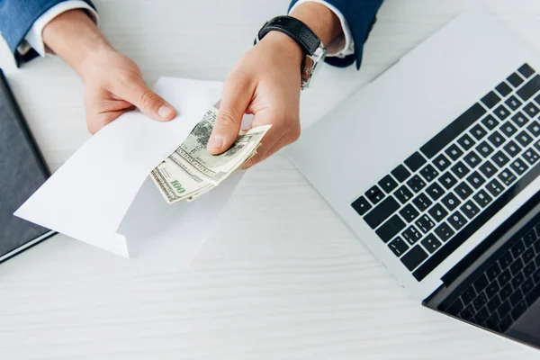 Vue du dessus de l'enveloppe d'un homme d'affaires tenant des billets en dollars près d'un ordinateur portable sur la table — Photo de stock