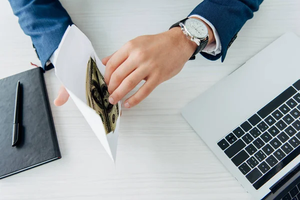 Обрізаний вид бізнесмена, що тримає конверт з доларовими банкнотами біля ноутбука на столі — стокове фото
