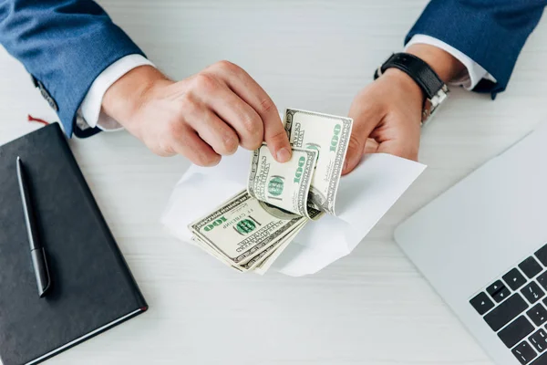 Cropped view of man holding envelope with dollar banknotes near laptop on table — Stock Photo