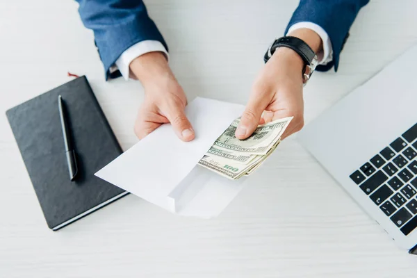 Вид сверху на человека, держащего конверт с деньгами возле ноутбука на столе — стоковое фото