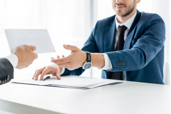 Vue recadrée de l'homme d'affaires donnant enveloppe avec de l'argent au partenaire d'affaires près du bureau — Photo de stock