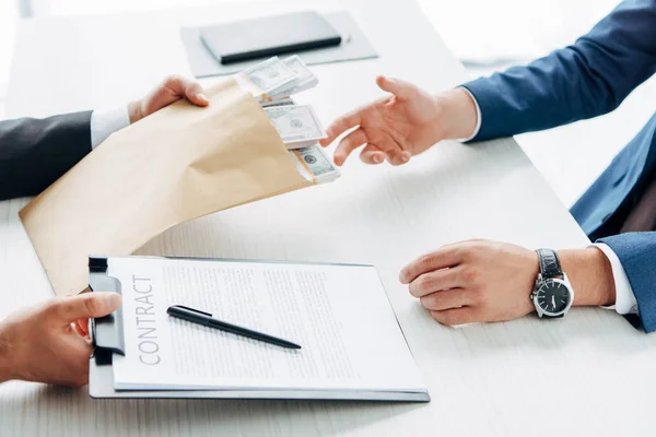 Visão cortada de homem tomando envelope com dinheiro perto de parceiro de negócios segurando área de transferência e contrato — Fotografia de Stock