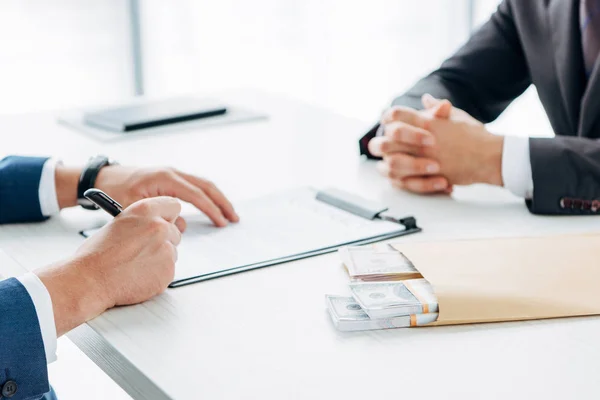 Selektiver Fokus des Geschäftsmannes unterschreibt Dokument in der Nähe Umschlag mit Bestechung und Geschäftspartner — Stockfoto