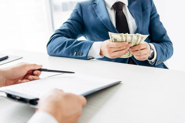 Vista recortada del hombre sujetando portapapeles y bolígrafo cerca de socio de negocios sosteniendo dinero - foto de stock