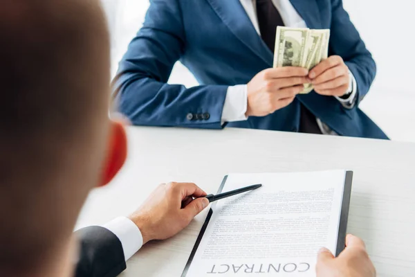 Selective focus of man holding clipboard and contract with pen near business partner holding money — Stock Photo