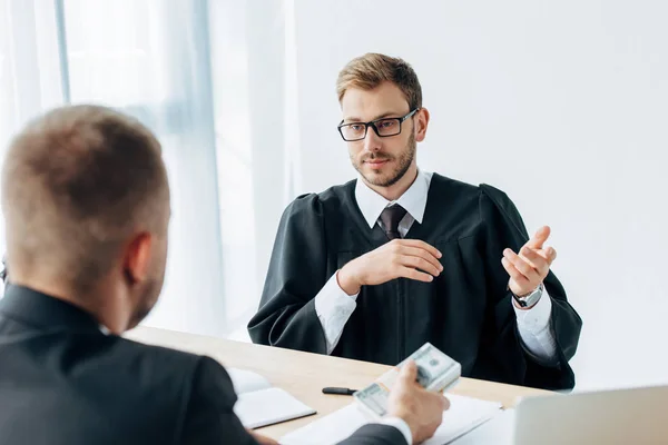 Selektiver Fokus eines gutaussehenden Richters mit Brille, der gestikuliert und einen Mann mit Dollarnoten ansieht — Stockfoto