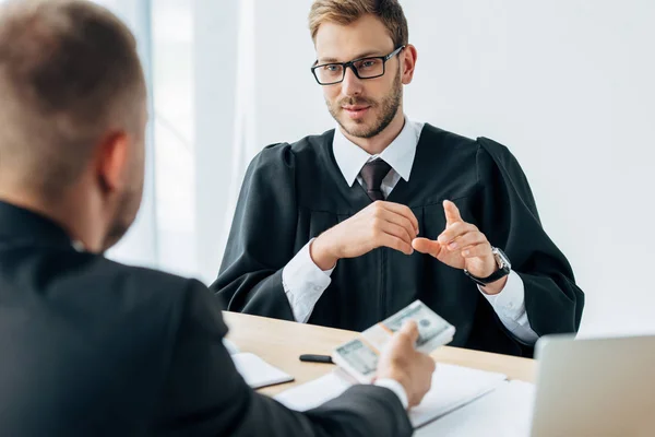 Selective focus of happy judge in glasses gesturing and looking at man holding dollar banknotes — Stock Photo