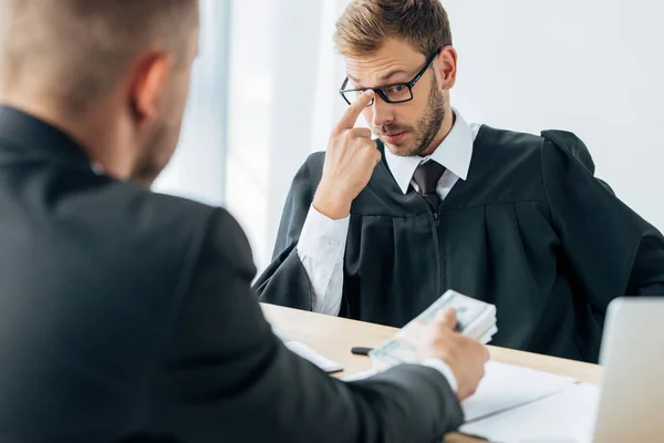 Messa a fuoco selettiva di bel giudice toccando occhiali e guardando banconote in dollari in mano sull'uomo — Foto stock