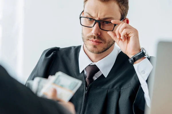 Enfoque selectivo de guapo juez tocando gafas y mirando dinero mano del hombre - foto de stock