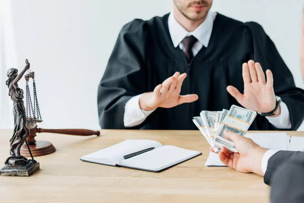 Enfoque selectivo del hombre dando dinero para juzgar sentado y gesticulando cerca de mazo de madera y estatuilla de la justicia - foto de stock