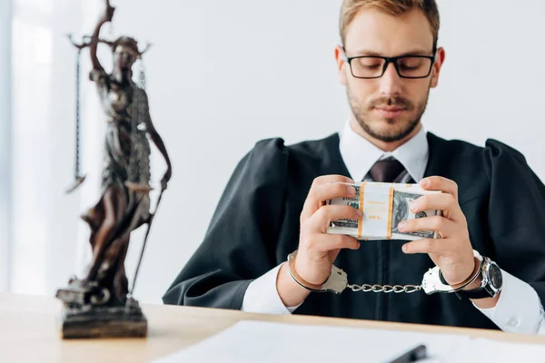 Enfoque selectivo de guapo juez esposado en vasos con billetes de dólar cerca de estatuilla de la justicia - foto de stock