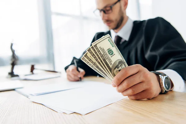 Selective focus judge in glasses holding dollar banknotes while writing in office — Stock Photo