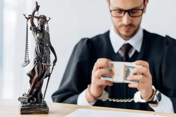 Foco seletivo da estatueta da justiça perto de belo juiz algemado em óculos segurando notas de dólar — Fotografia de Stock