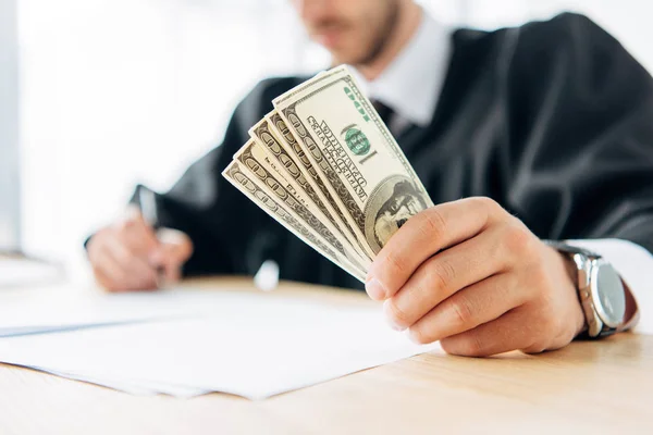 Selective focus of judge holding dollar banknotes and writing in office — Stock Photo