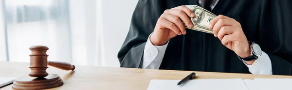 Panoramic shot of judge holding money near wooden gavel on table — Stock Photo