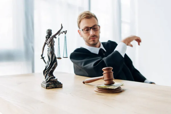 Selective focus of gavel with cash and statuette of justice near judge in glasses — Stock Photo