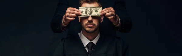 Panoramic shot of man standing and covering face of judge with dollar banknotes on black — Stock Photo