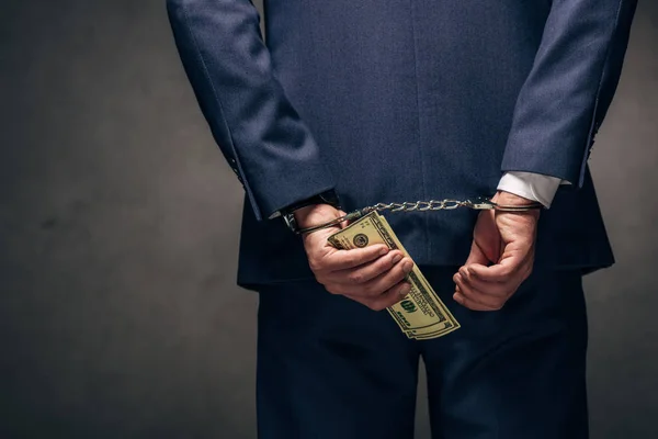 Cropped view of handcuffed businessman in suit holding dollar banknotes on grey — Stock Photo