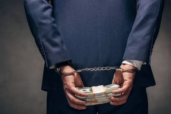 Обрізаний вид в наручниках бізнесмена в костюмі тримає хабар на сірому — стокове фото