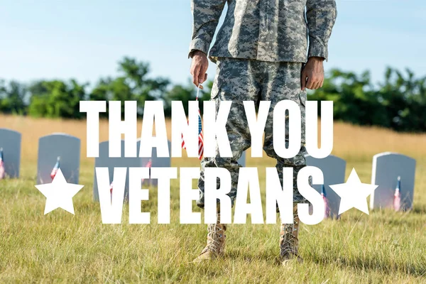 Cropped view of soldier in camouflage uniform holding american flag and standing in graveyard with thank you veterans illustration — Stock Photo