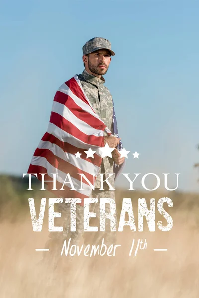 Selective focus of man in camouflage uniform and cap holding american flag in golden field with thank you veterans illustration — Stock Photo