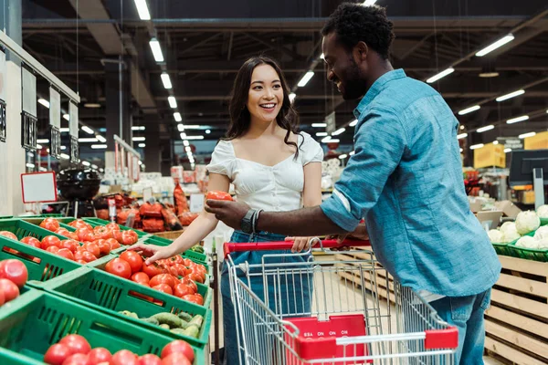 Felice ragazza asiatica e l'uomo africano americano in piedi vicino ai pomodori nel supermercato — Foto stock