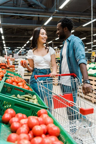 Foyer sélectif de fille asiatique heureuse et homme afro-américain debout près de tomates fraîches au supermarché — Photo de stock